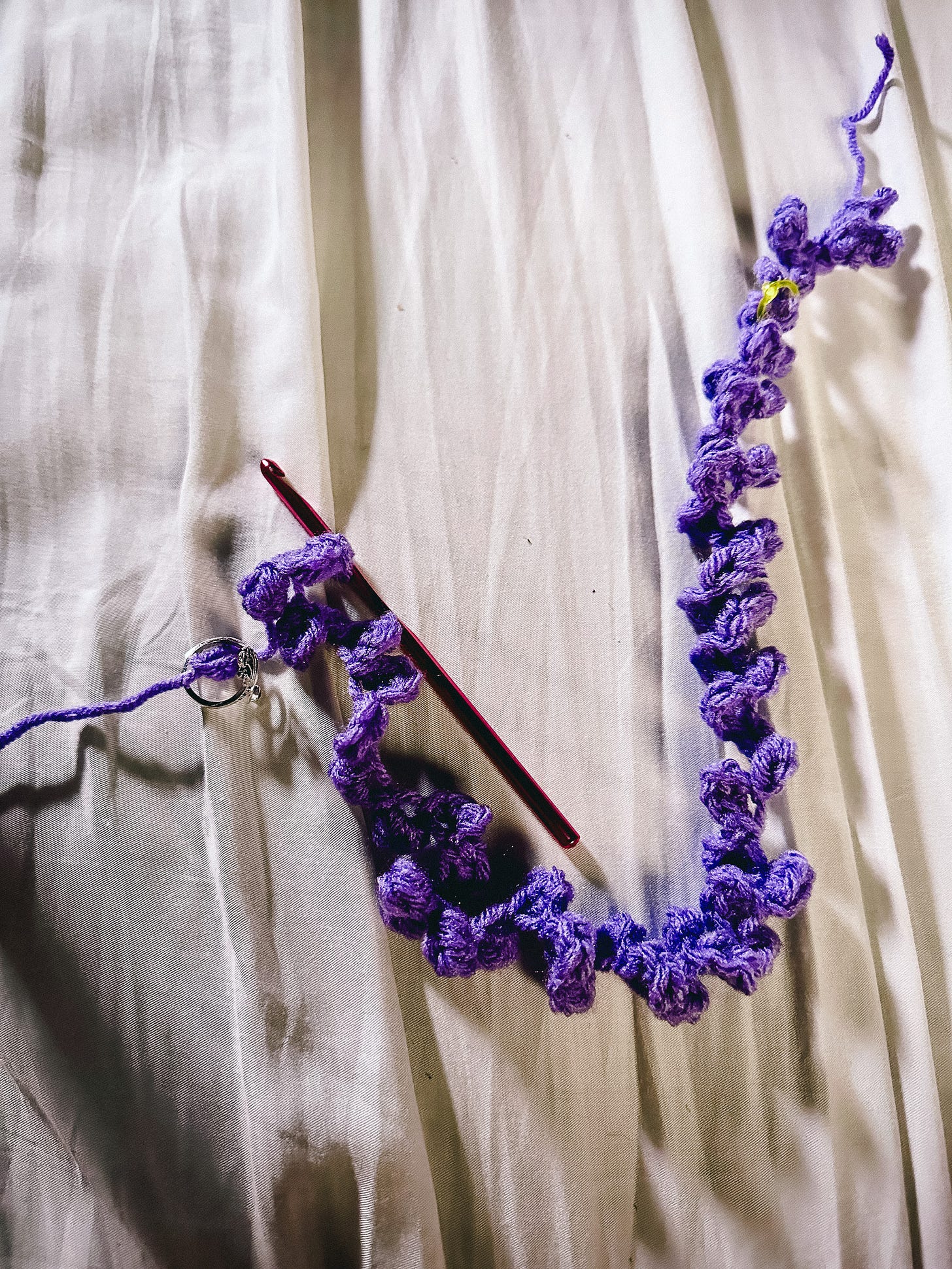 a string of purple yarn in the process of being crocheted into what looks like a bunch of knots, laying on a white bedsheet next to a crochet hook