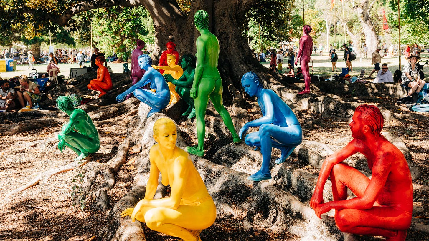 Nude performers painted bright solid colors, yellow, red, green and blue, in a public park.