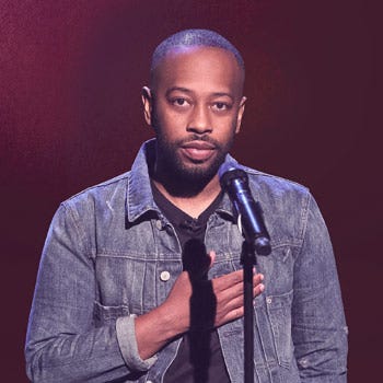 Picture of poet Rudy Francisco, standing behind a microphone, wearing a blue denim jacket holding his right hand over this heart.