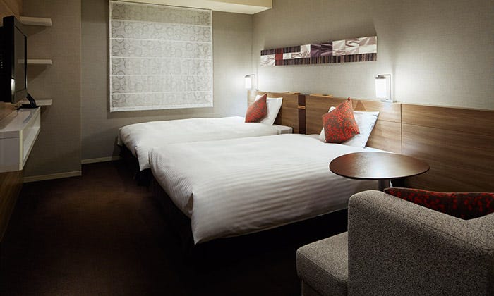 First Photo: Accessible twin room at Mitsui Garden Hotel Sapporo with two neatly made beds, soft lighting, and warm wood accents. The space includes a small table and armchair, creating a comfortable and modern atmosphere. Second Photo: Accessible bathroom at Mitsui Garden Hotel Sapporo featuring a toilet with support rail, roll-under sink, and bathtub with multiple grab bars. The design includes warm wood paneling and white tile for a clean, modern look. 