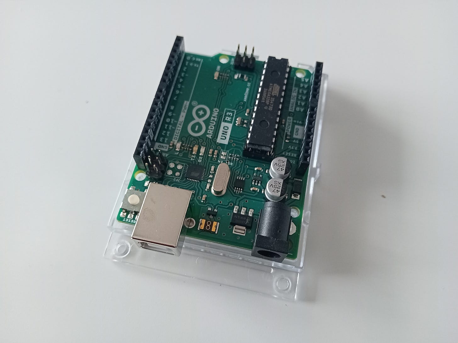 Arduino Uno Rev3 - płytka elektroniczna, na której szczegółowo widać różne układy elektroniczne m. in.: mikrokontroler ATmega, rezystory, kondensatory, diody, przycisk. Widać też złącza umożliwiające wpięcie przewodów sygnałowych Arduino Uno Rev3 - płytka elektroniczna, na której szczegółowo widać różne układy elektroniczne m. in.: mikrokontroler ATmega, rezystory, kondensatory, diody, przycisk. Widać też złącza umożliwiające wpięcie przewodów sygnałowych