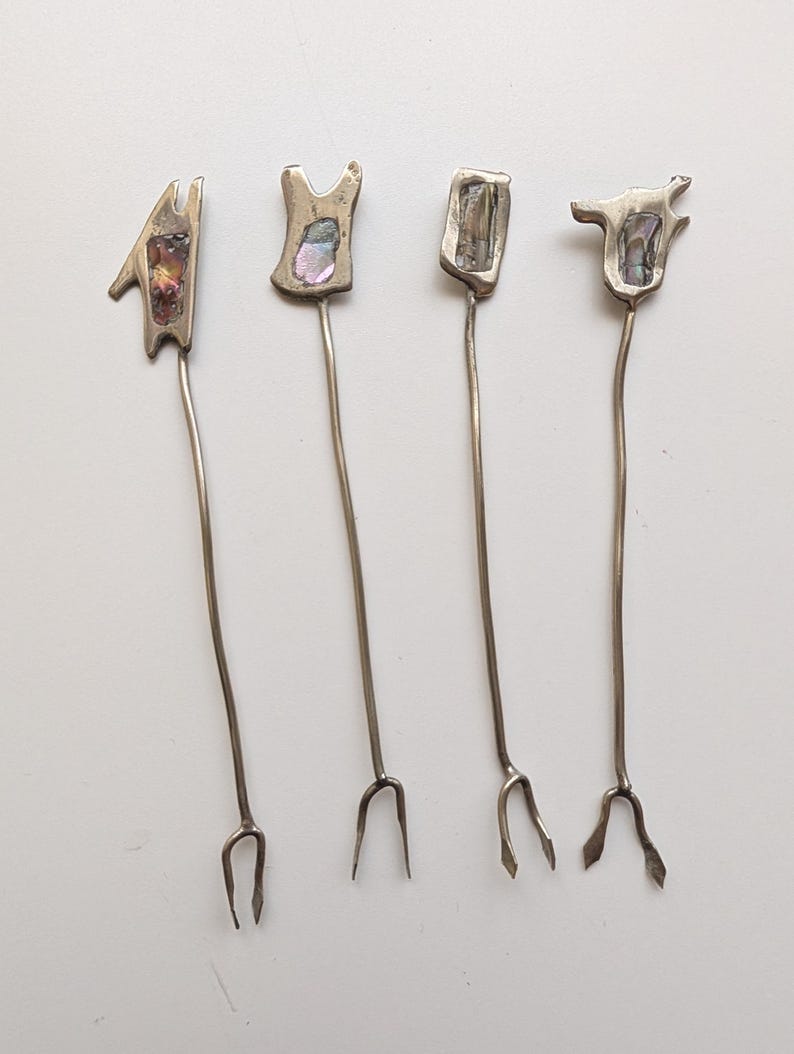 May include: Four silver-toned metal appetizer forks with long, thin stems and two-pronged tips. Each fork features a unique, irregular head shape inlaid with a small, iridescent abalone shell piece that displays shifting colors of purple, green, and blue.