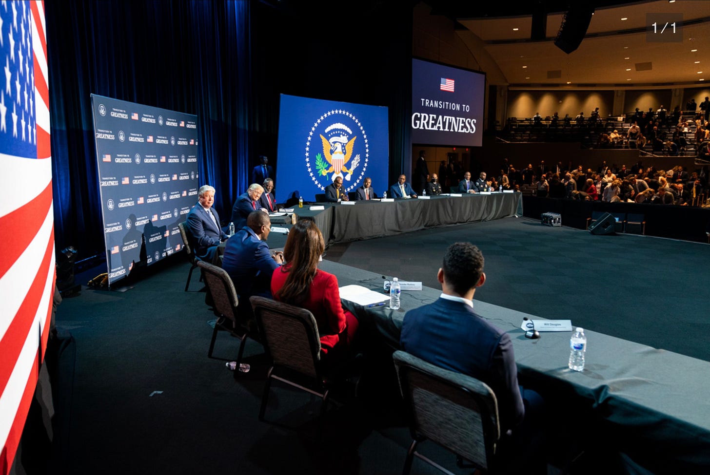 A large audience hall / church with Trump, Robert Morris and other speakers on stage. American flags the seal of the presidency and "Transition to Greatness" are on stadium-sized TV screens. Crowd is offset due to covid-19 distancing. A large audience hall / church with Trump, Robert Morris and other speakers on stage. American flags the seal of the presidency and "Transition to Greatness" are on stadium-sized TV screens. Crowd is offset due to covid-19 distancing.