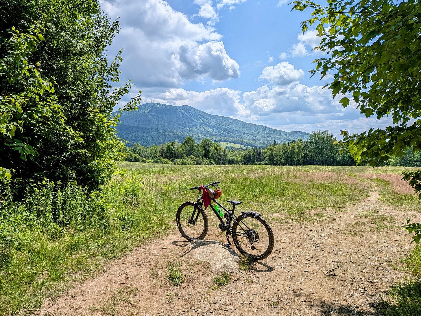 A black full-suspension mountain bike resting on a dirt trail at the edge of a grassy meadow. Burke Mountain rises in the background with visible ski trails cutting down its slopes. The scene is framed by lush green trees on either side under a bright blue sky with white cumulus clouds. A black full-suspension mountain bike resting on a dirt trail at the edge of a grassy meadow. Burke Mountain rises in the background with visible ski trails cutting down its slopes. The scene is framed by lush green trees on either side under a bright blue sky with white cumulus clouds.