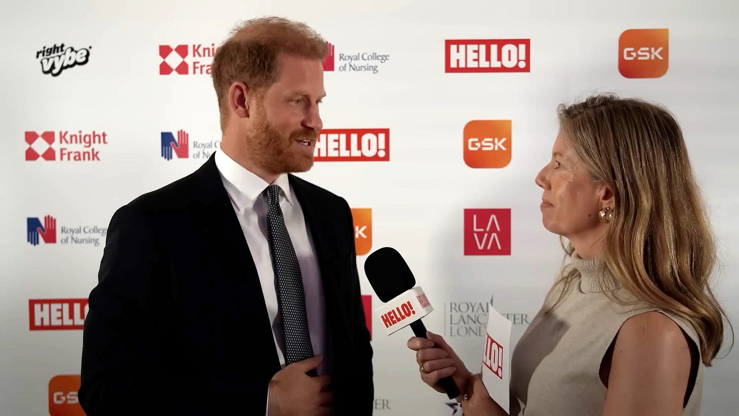Women in short sleeved cream top holding a microphone while interviewing a suited Prince Harry with both standing against a white backdrop with corporate logos Women in short sleeved cream top holding a microphone while interviewing a suited Prince Harry with both standing against a white backdrop with corporate logos