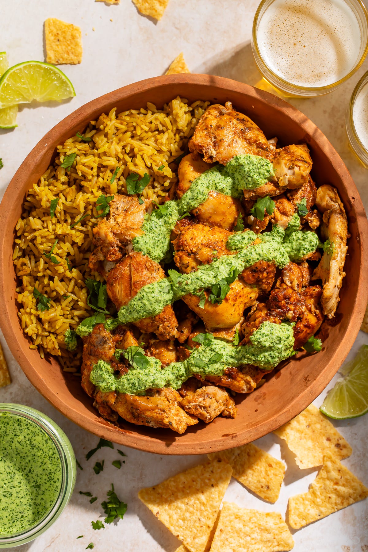 A terra cotta bowl filled with yellow rice, topped with slow cooker Peruvian chicken and drizzled with aji verde (green sauce). Surrounded by chips, cups of beer, and lime wedges