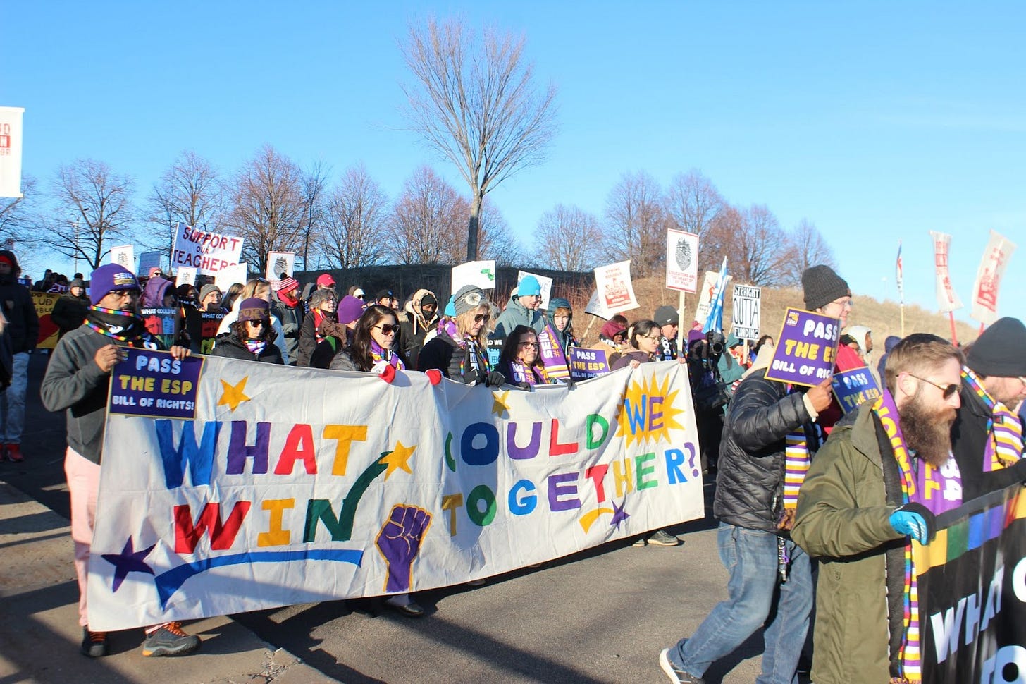 a crowd of people march in the street carrying picket signs and a banner reading "What could we win together?" a crowd of people march in the street carrying picket signs and a banner reading "What could we win together?"
