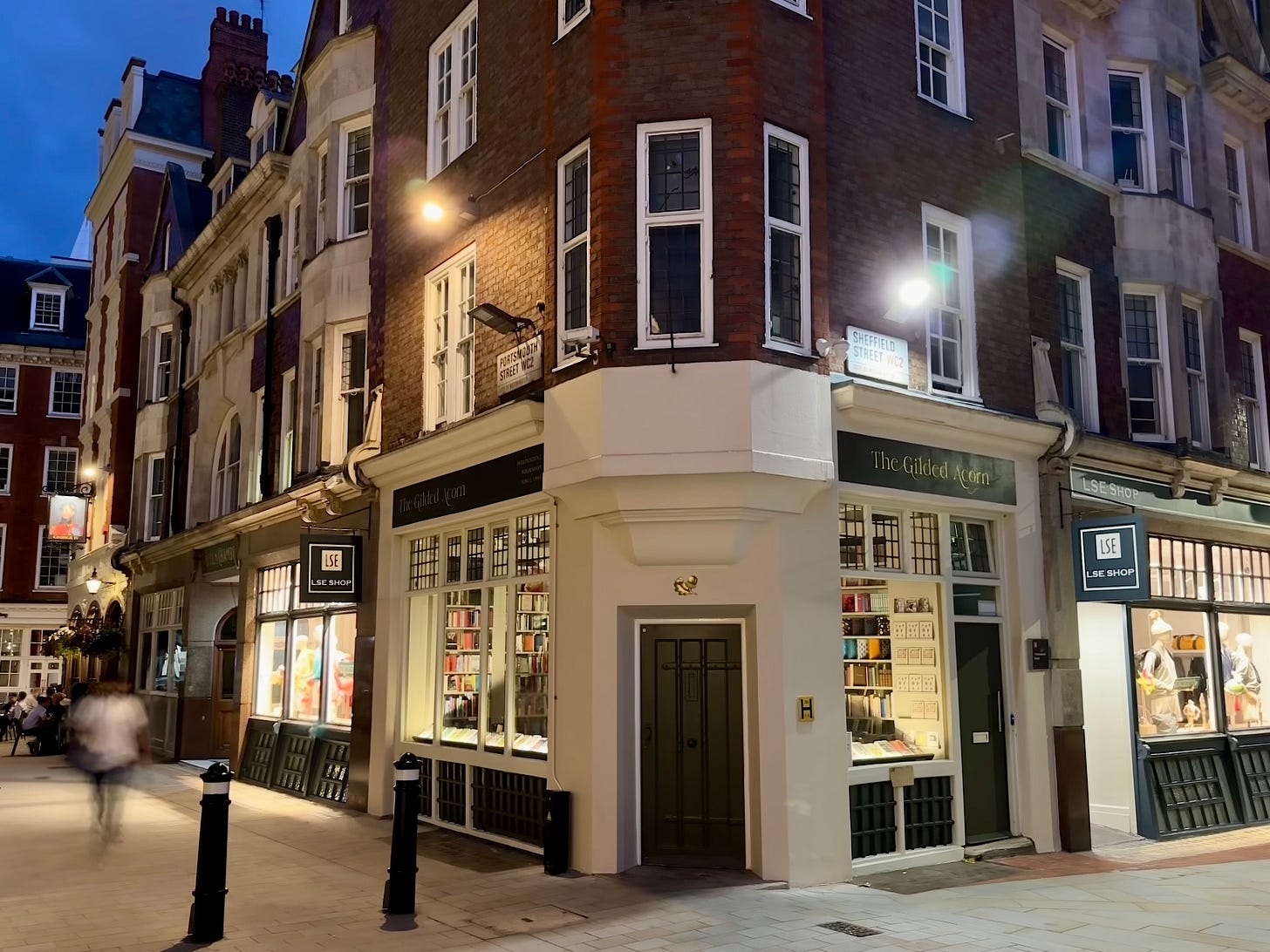 A photo taken near dusk on a balmy summer evening of the independent bookshop called The Gilded Acorn, near the London School of Economics. House in a beautifully restored corner unit of an Edwardian redbrick terrace, the frontage of the bookshop looks out over a well kept courtyard.