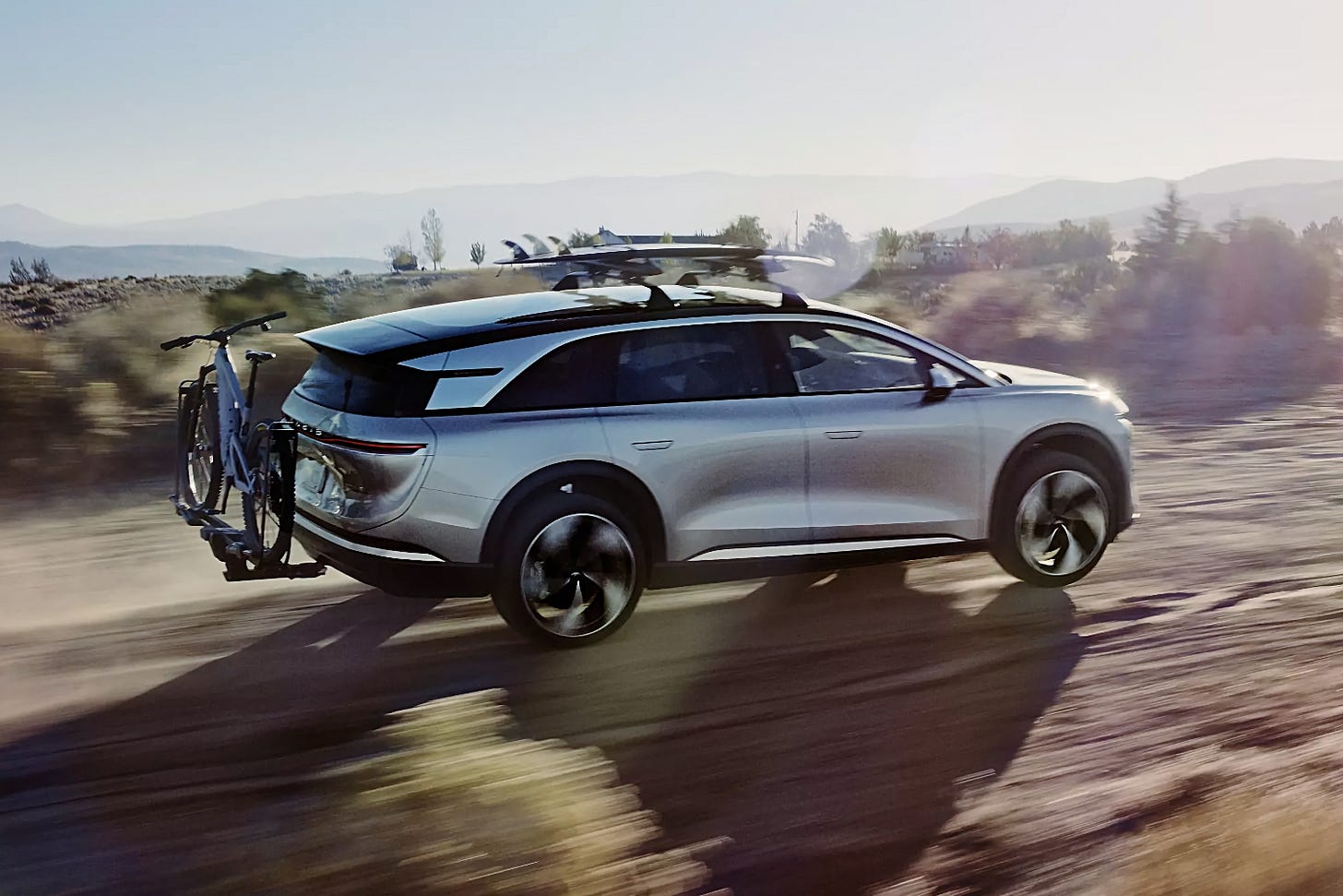 A silver Lucid Gravity is driving off-road in a desert with surfboards on the roof and a mountain bike in a rack on the back.