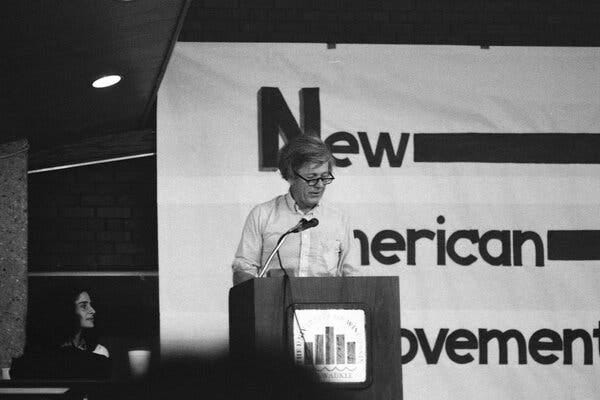 A man in a button-down shirt and glasses speaks from a podium. He is partly blocking from view a banner that reads, “New American Movement.”