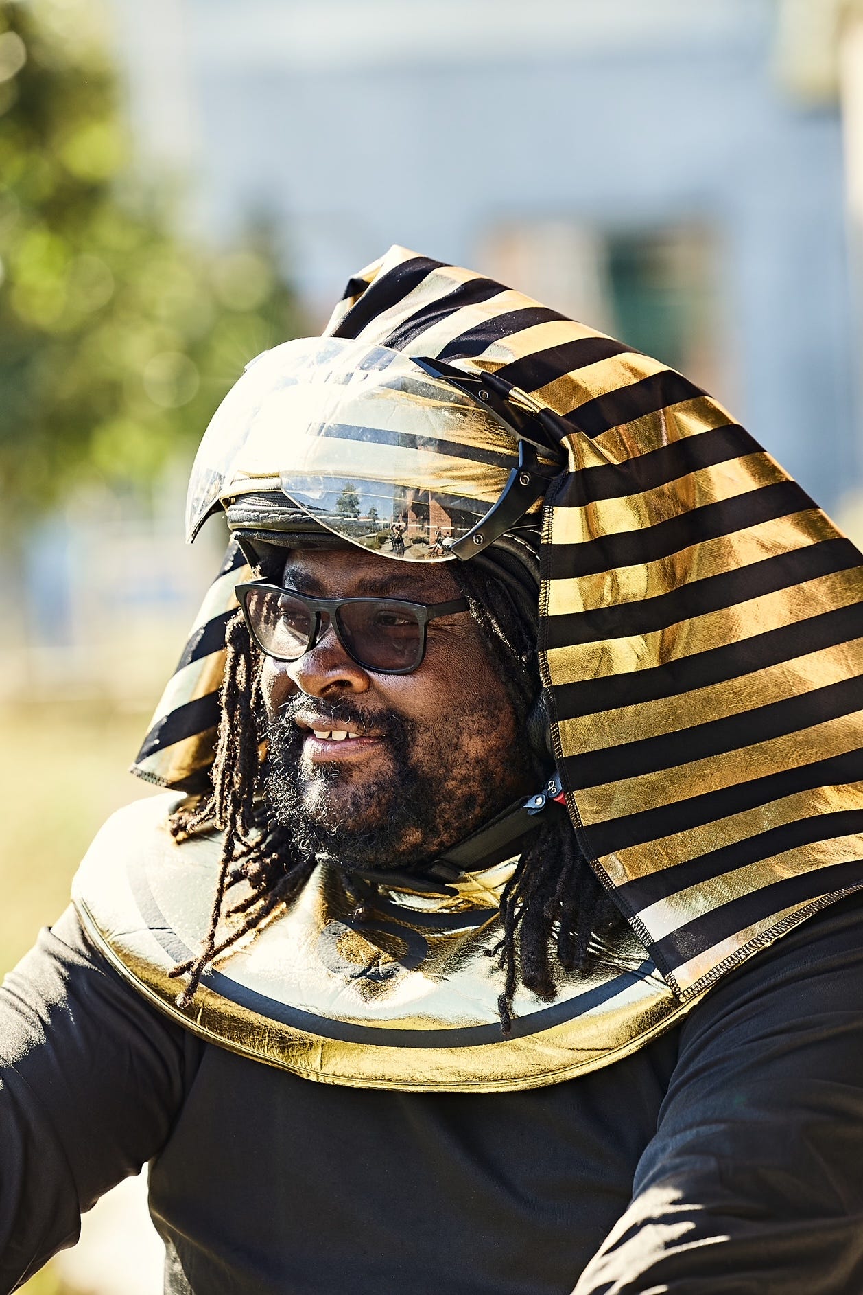 Portrait of FatbikeHero wearing a black and gold striped Egyptian-style headdress and gold collar, with goggles and glasses, seated outdoors in bright daylight.
