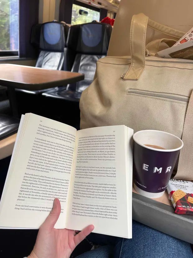Hand holding a paperback book open next to a paper cup of tea and packet of shortbread biscuits. Travel bag and train car in the background.