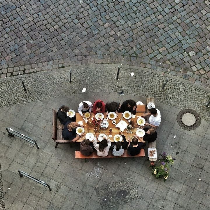 This may contain: a group of people sitting around a wooden table eating food on top of a sidewalk