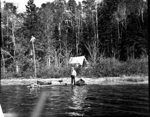 Image of a horse and wagon on a woodland road, a guide is working on a tree blocking the way; a campsite of Clifford Lake, Maine