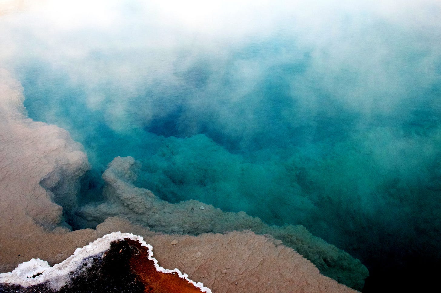 A thermal pool in Yellowstone National Park features a salt-rimmed red earth edge dropping off into deep turquoise waters shrouded in steam.