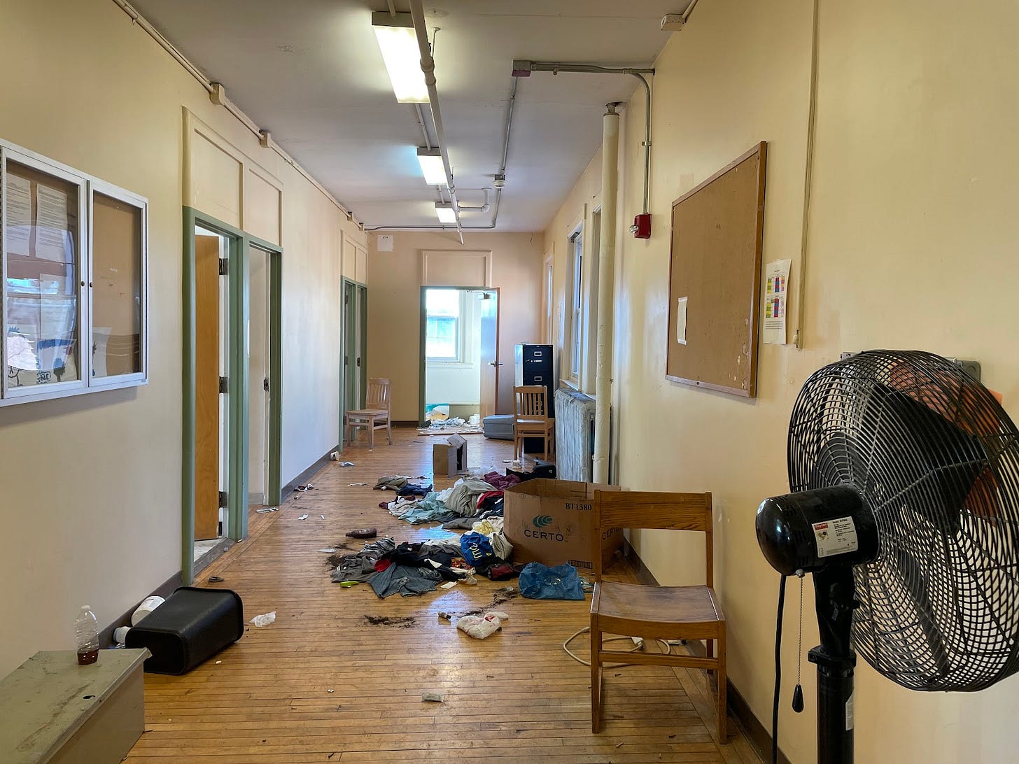 A view of a hallway in the former Long Island public health campus. Chairs and a mattress are leaned against the wall, and clothes are on the floor next to a cardboard box. A small trash can is knocked down. A plastic bottle with a brown liquid still sits on a table. A view of a hallway in the former Long Island public health campus. Chairs and a mattress are leaned against the wall, and clothes are on the floor next to a cardboard box. A small trash can is knocked down. A plastic bottle with a brown liquid still sits on a table.