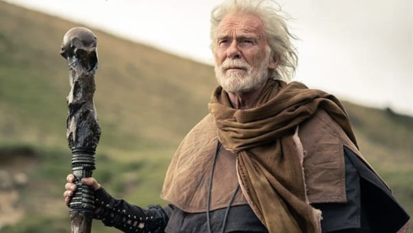 This image shows the character of Ko Sharmus (played by Ian McElhinney) standing near a hill; holding a large, ornately carved staff in his right hand; and looking offscreen (at his lonely, deserted encampment). This image shows the character of Ko Sharmus (played by Ian McElhinney) standing near a hill; holding a large, ornately carved staff in his right hand; and looking offscreen (at his lonely, deserted encampment).