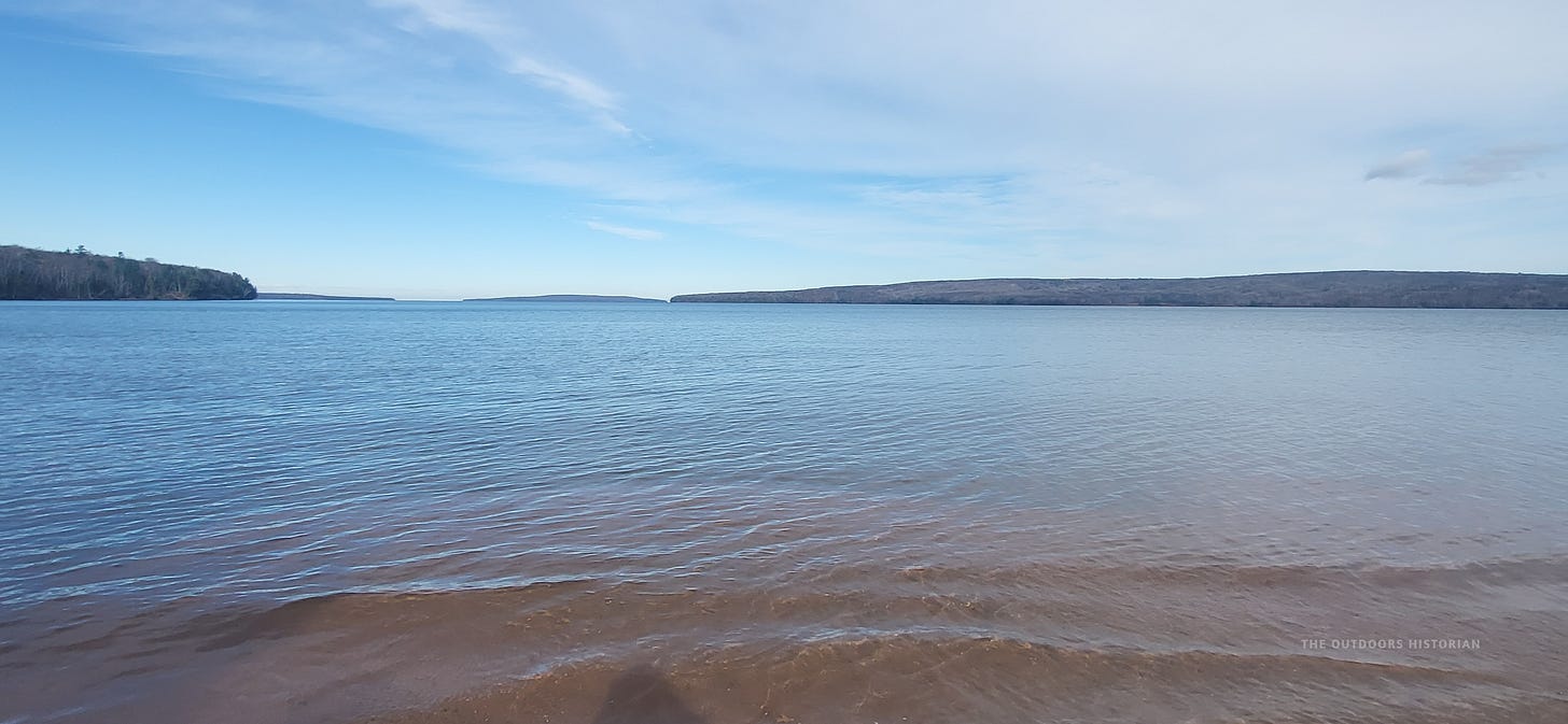 A wide-angle landscape photograph of Frog Bay on a calm, sunny day looking northwest. The clear water of Lake Superior ripples gently in the foreground. Across the water, the green, forested profile of Oak Island sits on the horizon, with the faint silhouette of Bear Island visible in the far distance under a bright sky.