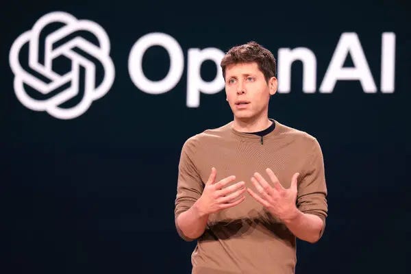 A speaker stands on stage in front of the OpenAI logo, gesturing during a presentation about artificial intelligence innovation and company direction. A speaker stands on stage in front of the OpenAI logo, gesturing during a presentation about artificial intelligence innovation and company direction.