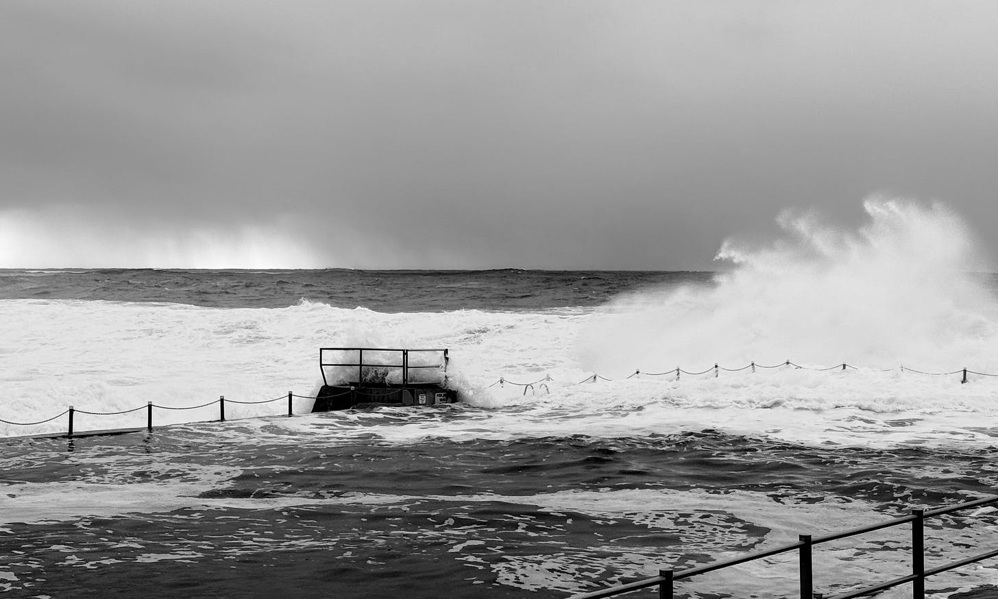 Black and white image of big surf crashing into an ocean pool barrier with salt spray.