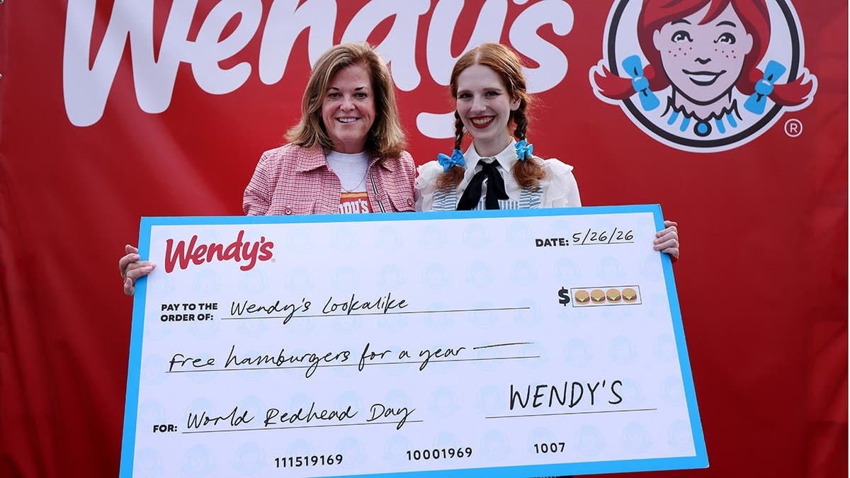 A Wendy impersonator stands with another woman behind a large check written out for free hamburgers. A Wendy impersonator stands with another woman behind a large check written out for free hamburgers.