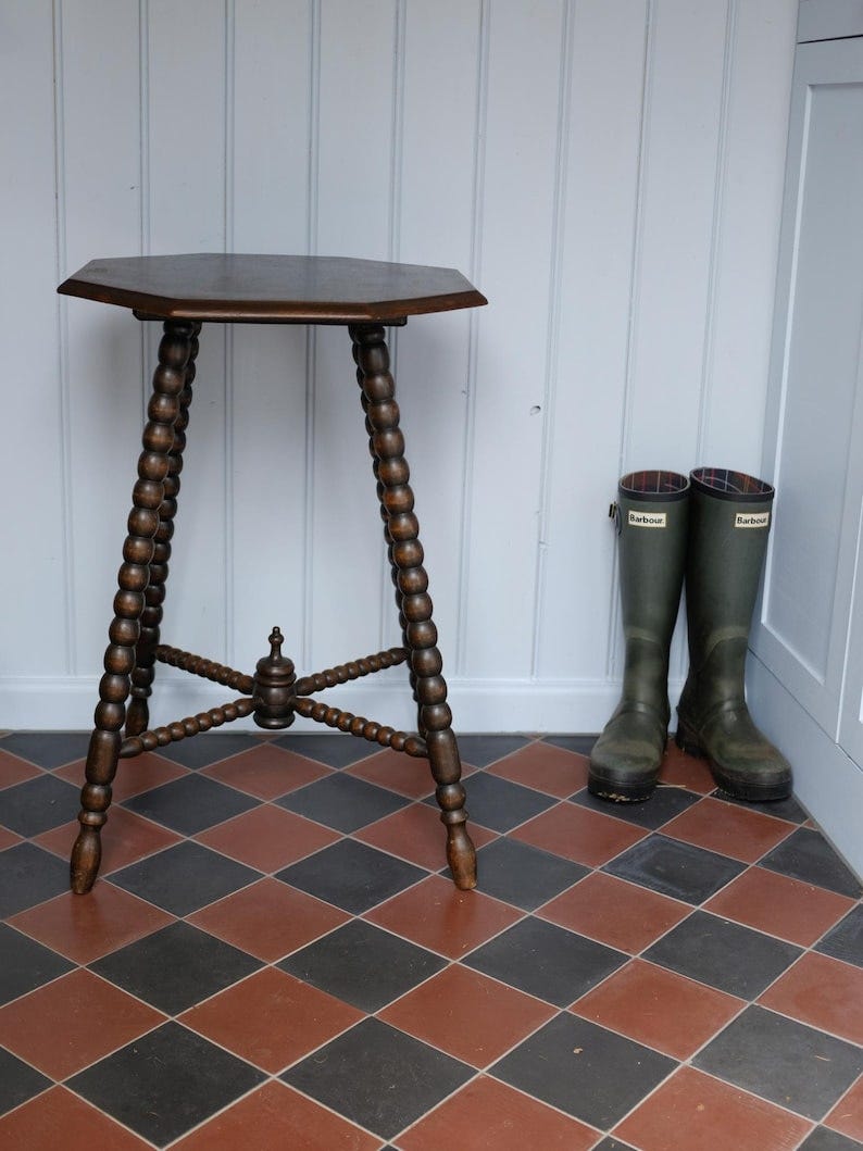 May include: A dark brown, antique wooden side table with an octagonal top and intricately carved, bobbin-turned legs. The table stands on a checkered floor, with a pair of green rain boots resting against the wall. The table is approximately 28 inches tall.
