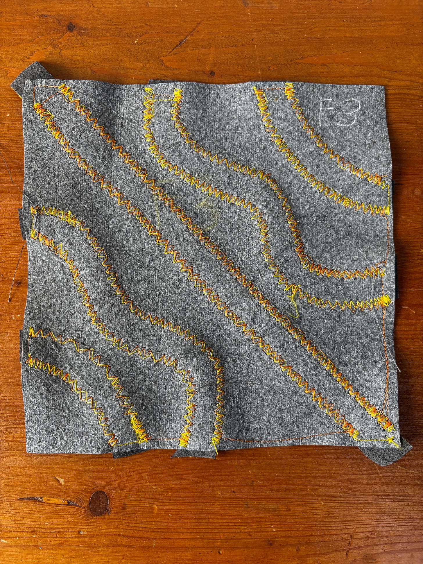A 20 centimetre by 20 centimetre piece of grey fabric on a wood tabletop. There are wavy lines of zigzag stitching in yellow and orange thread.