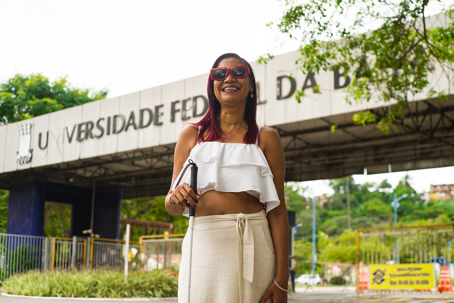 Imagem em ambiente externo, durante o dia. Em primeiro plano, está Cristiane de Jesus, mulher adulta, sorridente, de pele negra, em pé, voltada para a câmera. Ela usa óculos escuros com armação vermelha, cabelo liso na altura dos ombros, tingido em tom avermelhado. Veste uma blusa branca sem mangas, com babados na parte superior, deixando o abdômen à mostra, e uma saia longa clara, de cintura alta, com amarração frontal.  Na mão direita, ela segura uma bengala branca, posicionada à frente do corpo.   Ao fundo, aparece a entrada de uma universidade, com uma grande estrutura de concreto e uma placa onde se lê “Universidade Federal da Bahia”. Há árvores com folhagem verde ao redor, grades metálicas e cones de sinalização mais ao fundo, indicando área urbana e institucional. O céu está claro, sugerindo clima ensolarado.
