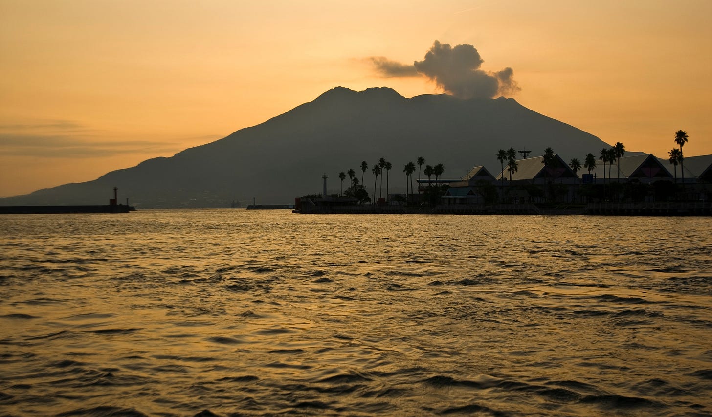 Silhouette of the smoking Sakurajima Volcano rising over the water at sunset, lined with palm trees along the shore.