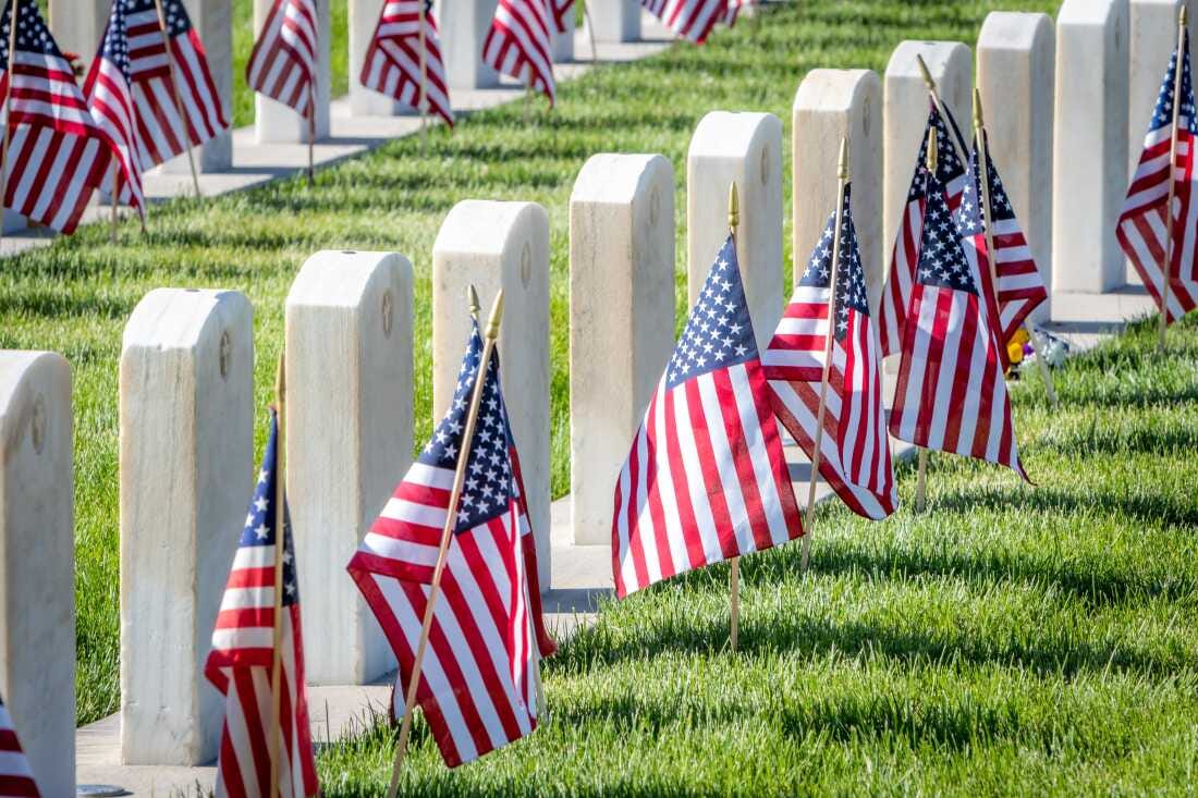 This photo shows two rows of military headstones, each with an American flag staked into the grassy green ground in front of it. This photo shows two rows of military headstones, each with an American flag staked into the grassy green ground in front of it.