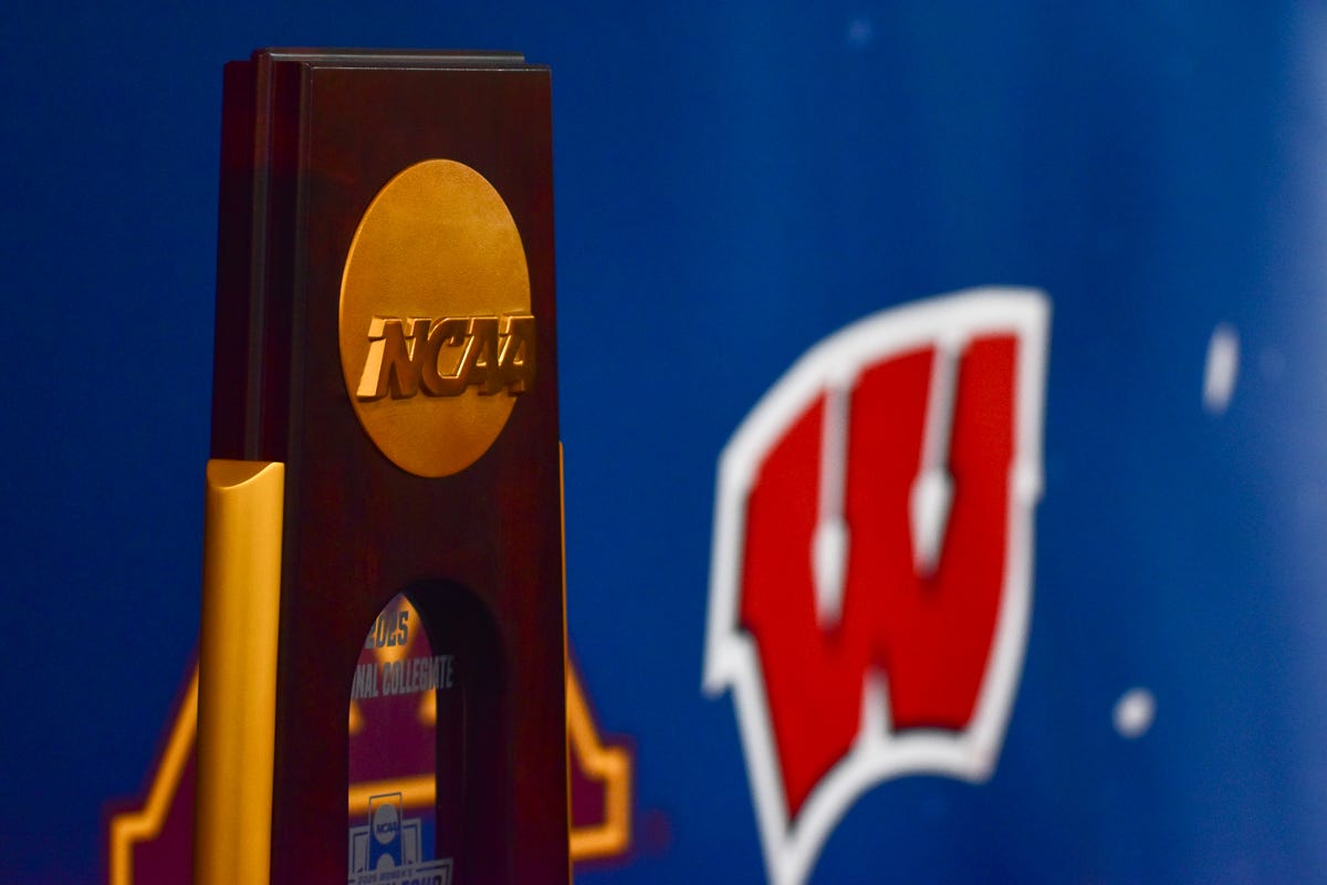 the ncaa tournament trophy sits in front of a banner where you can see the Wisconsin Badgers 'motion W' logo to the right