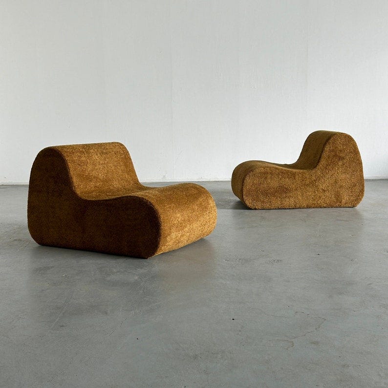 May include: Two brown, fuzzy, modern lounge chairs with rounded edges. The chairs are sitting on a gray concrete floor.