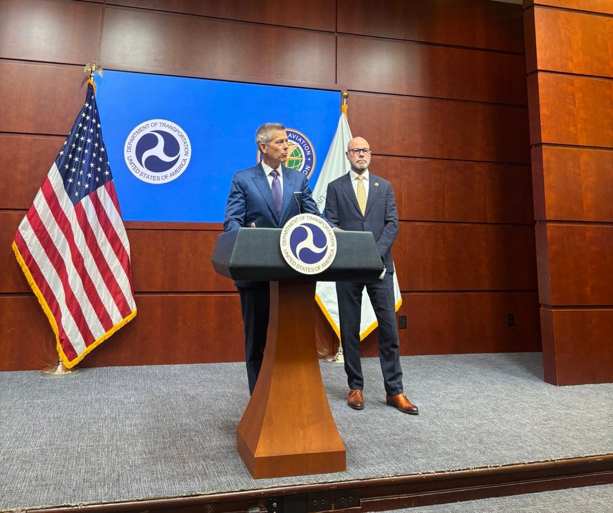 Two men in suits stand at a wooden podium with microphones in a wood-paneled room featuring American and blue flags with circular emblems on the backdrop behind them