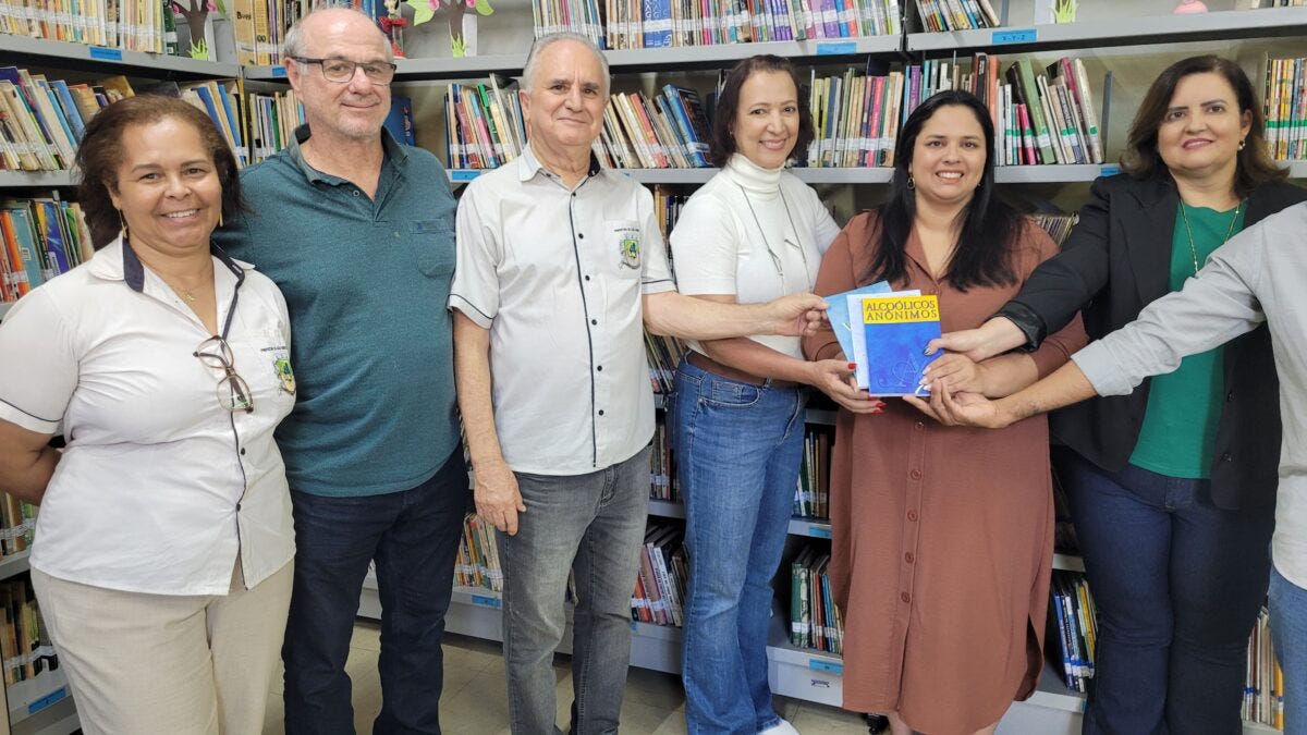#descrevemospravocê  A imagem mostra seis pessoas em pé, lado a lado, em frente a estantes repletas de livros em uma biblioteca. Elas estão sorrindo e posam para a foto enquanto seguram juntas um exemplar do livro “Alcoólicos Anônimos”, cuja capa é azul com a faixa superior amarela onde está escrito o título em letras azuis. Da esquerda para a direita, há uma mulher de pele parda com cabelos castanhos presos, vestindo camisa branca com detalhes pretos e calça clara, com óculos pendurados na gola; ao lado, um homem de pele clara, calvo e de óculos, usando camisa polo verde escura e calça jeans; o terceiro é um homem de pele clara, cabelo grisalho, usando camisa social clara com detalhes escuros e calça jeans; a quarta pessoa é uma mulher de pele clara, cabelo castanho escuro, usando blusa de manga longa branca e calça jeans; a quinta é uma mulher de pele clara e cabelo escuro, usando vestido marrom de manga longa, segurando o livro com as demais; por fim, à direita, está uma mulher de pele clara, cabelo castanho escuro, vestindo blazer preto, blusa verde e calça jeans. Um braço de outra pessoa aparece à direita, também segurando o livro. Ao fundo, nas estantes, há livros organizados com etiquetas coloridas, plantas decorativas e pequenos enfeites no topo, criando um ambiente de biblioteca pública acolhedor.