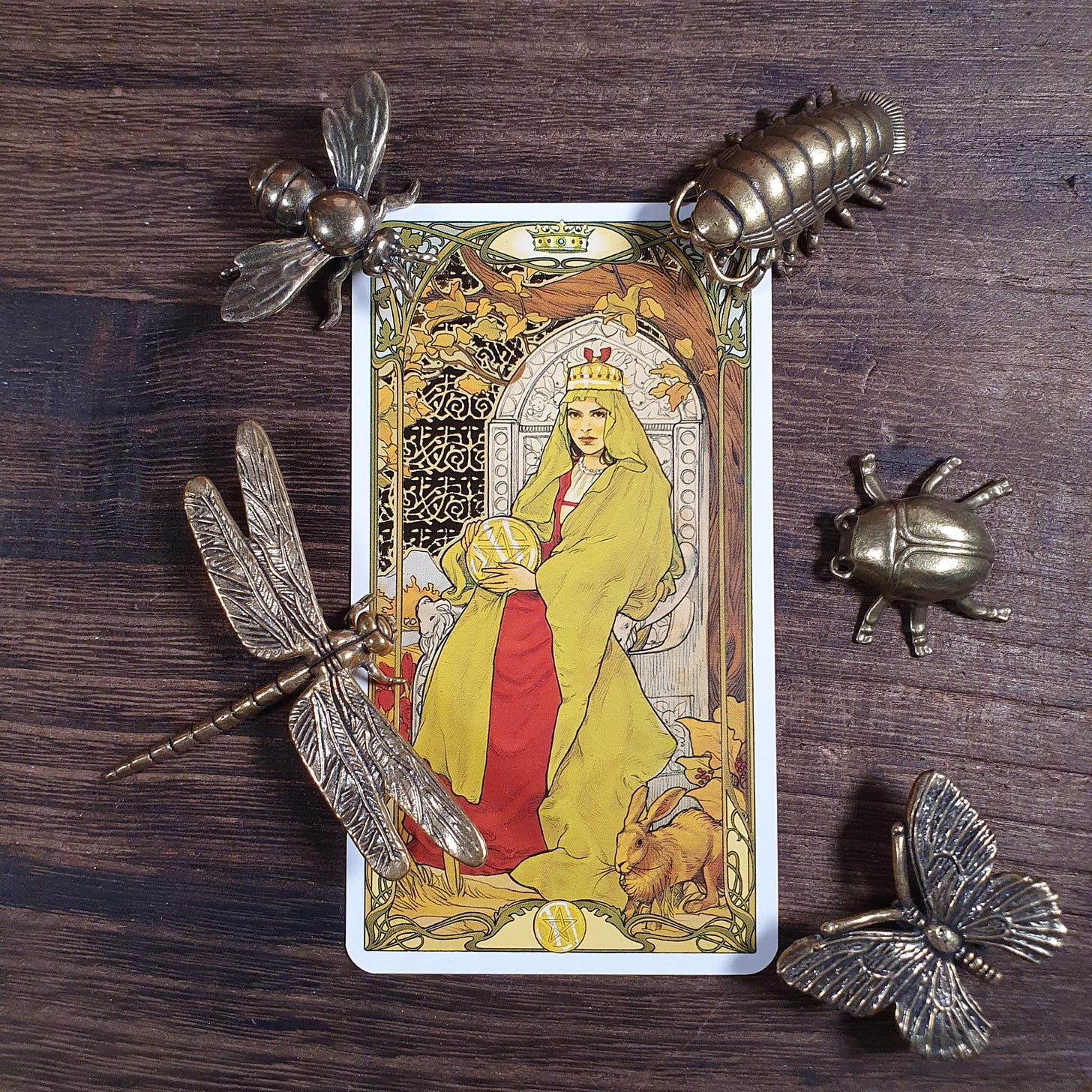 Queen of Pentacles card from the Golden Art Nouveau Tarot surrounded by brass insect ornaments including dragonfly and beetle on wooden surface, photographed for the Tarot DMs interview with Alice Slater by Finbarre Snarey.