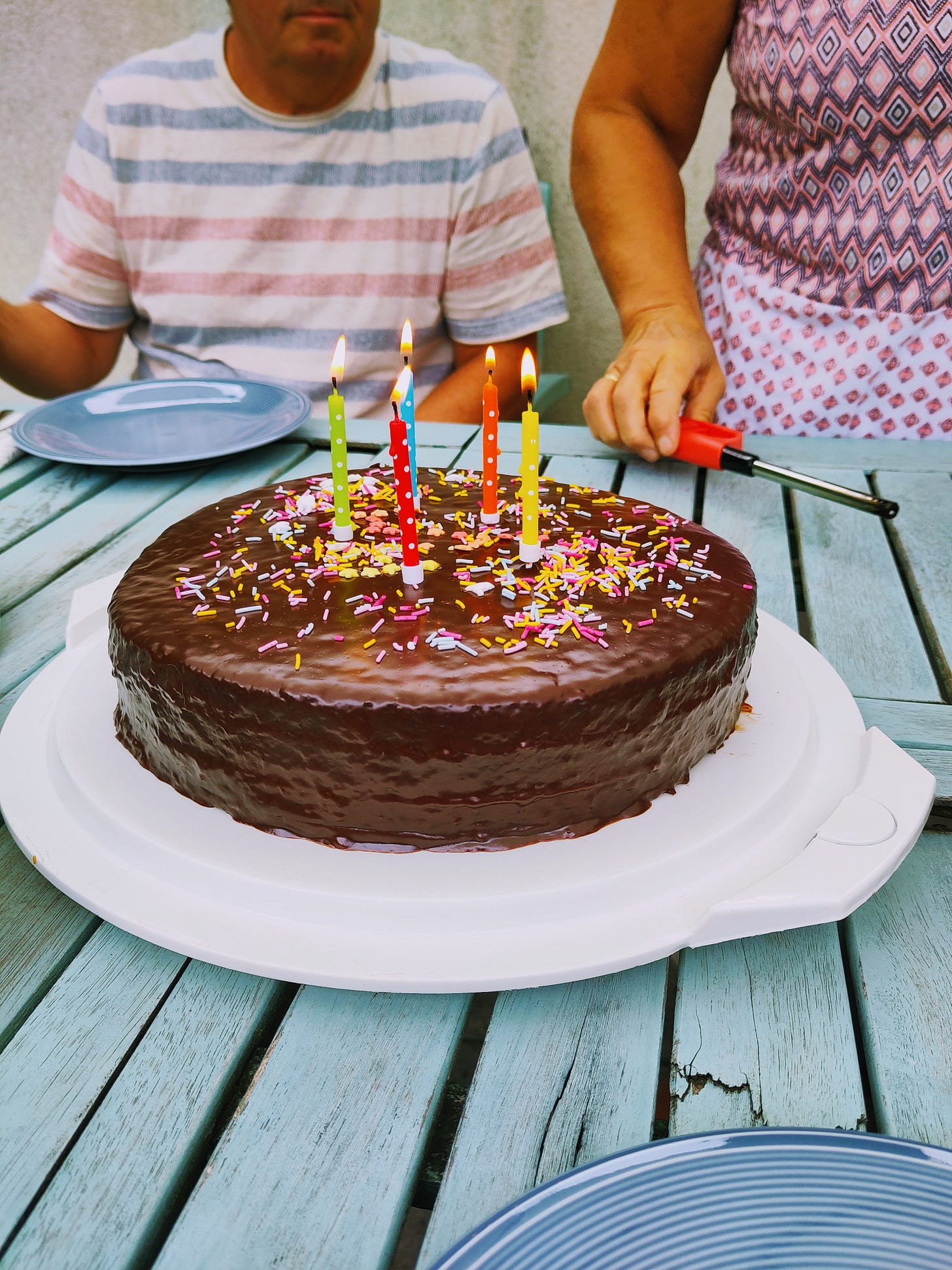 In the center of the photo is a round chocolate cake with five burning candles on them. In the background are a man and a woman wearing colorful shirts, but you cannot see their faces.