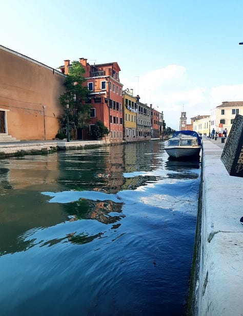 Image 1: A picturesque stone bridge crossing a Venetian canal at sunset, with people walking across and reflections of historic Italian architecture shimmering in the blue water. A small boat is docked on the left, and a terracotta building with white trim stands on the right. Photo taken in Venice, Italy. Image 2: Outdoor summer gathering in an Italian backyard, featuring a plastic cup with a pink cocktail in the foreground, palm trees, and a group of friends playing beer pong in the background. The sun is shining behind the trees, giving a relaxed Italian party vibe. Photo taken in Italy. Image 3: Classic Venetian canal scene with colorful historic buildings, a moored boat, and calm water reflecting the architecture. The canal stretches toward a distant clock tower under a bright blue sky. Photo taken in Venice, Italy. Image 4: Black-and-white photograph of Venetian canal houses and rippling water, highlighting the texture and timeless character of Italy’s iconic canal-side architecture. Taken in Venice, Italy. Image 5: Close-up of a gray cat perched on a stone ledge with lush Italian greenery behind, while another cat lounges on the pavement next to a blue car. The evening sky is partly cloudy, capturing a quiet residential moment in Italy.