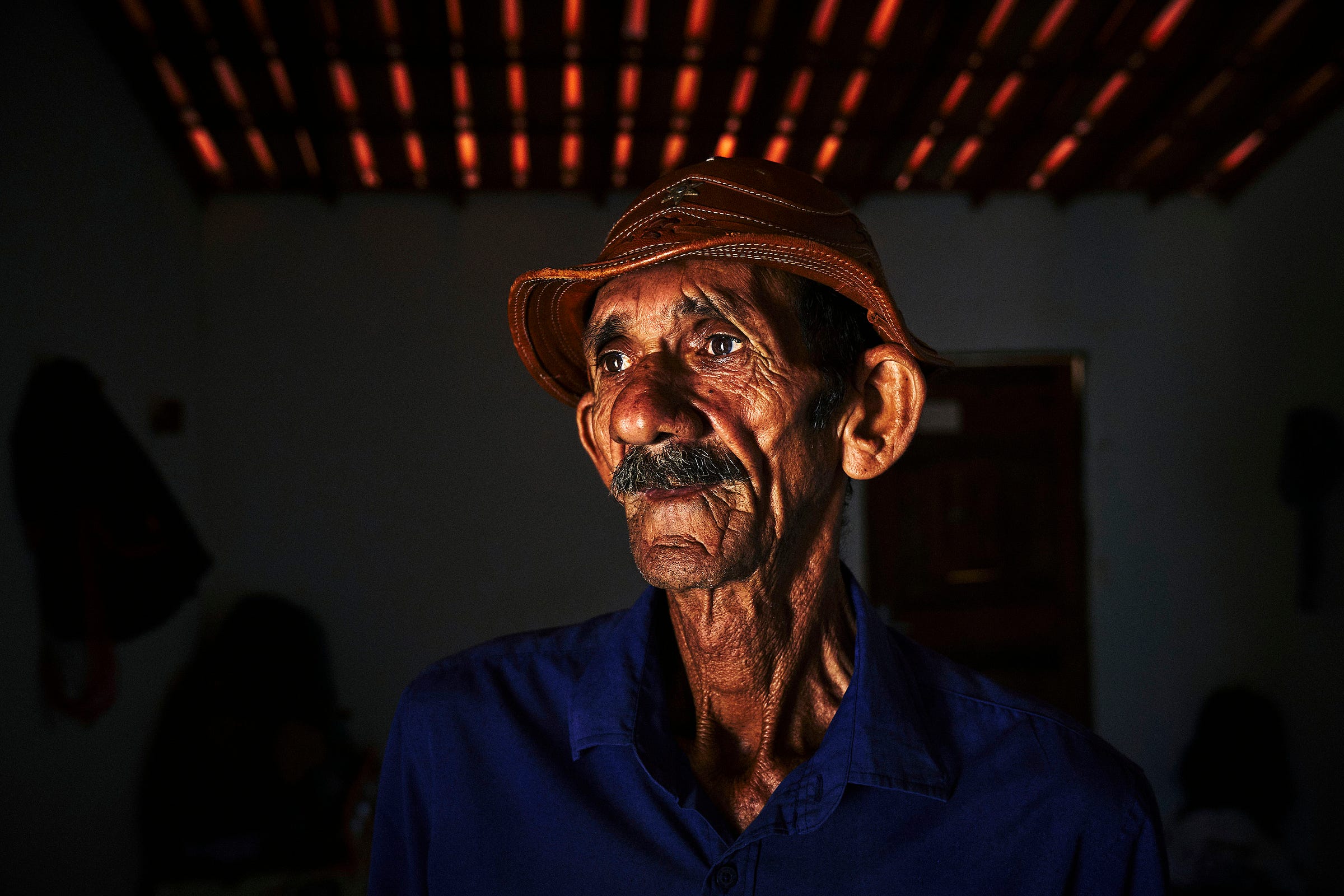 An older man with a weathered face and mustache, wearing a brown hat and blue shirt, stands indoors under a ceiling with slats, gazing thoughtfully into the distance. The background is dark and softly lit.