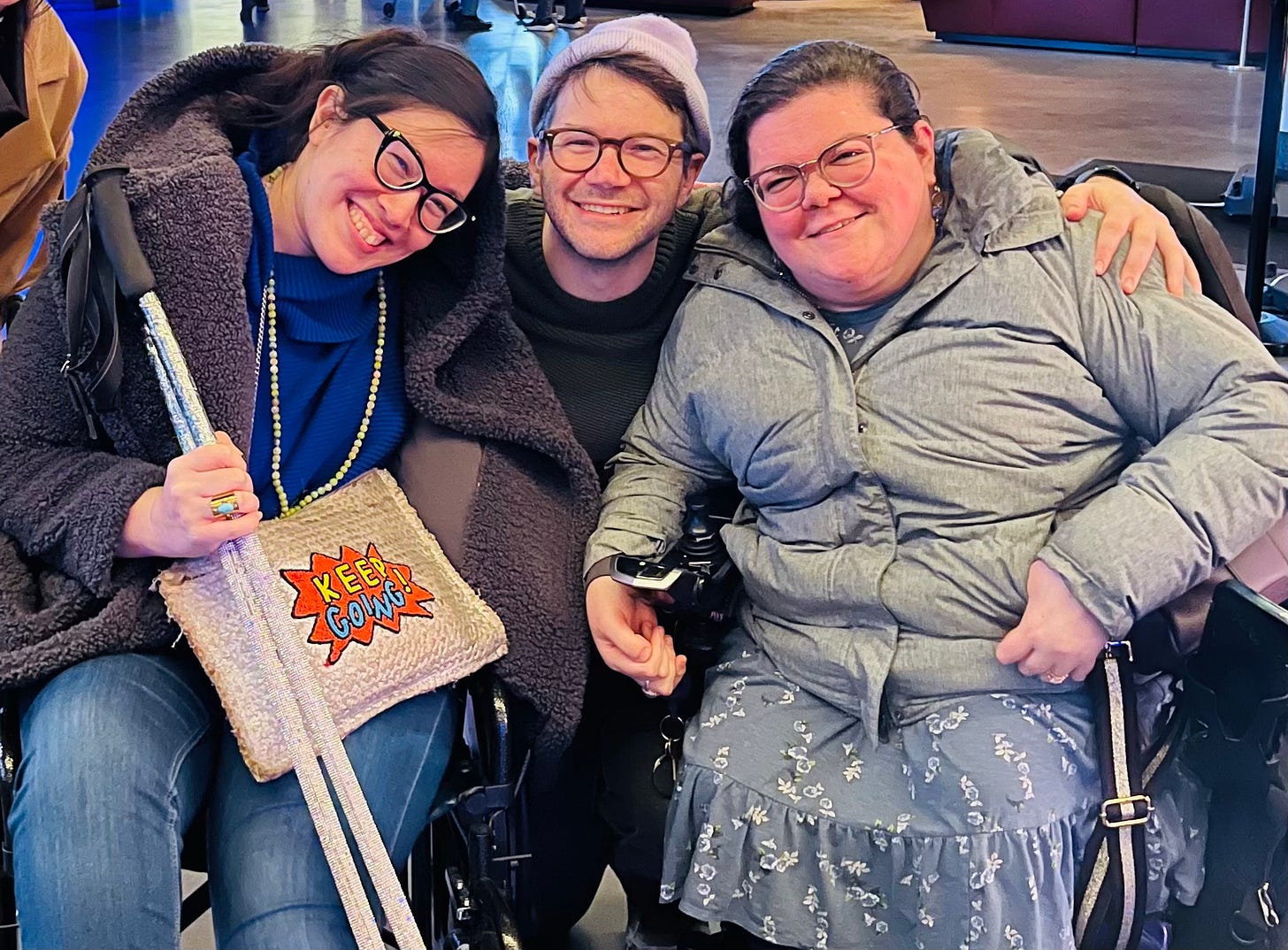 Xian, a joyful half-Asian woman, sitting in a wheelchair and holding the ski poles she uses to walk, alongside James Caverly, a white man smiling in glasses and lavender beanie, and me, a white woman, also sitting in my wheelchair. Xian, a joyful half-Asian woman, sitting in a wheelchair and holding the ski poles she uses to walk, alongside James Caverly, a white man smiling in glasses and lavender beanie, and me, a white woman, also sitting in my wheelchair.