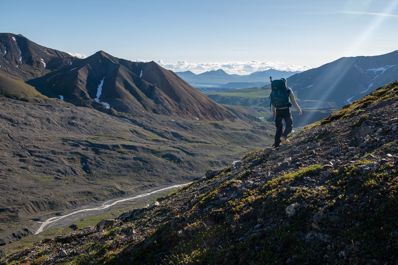 A hiker in a teal backpack, holding a trekking pole, climbs a steep slope of rock and low green tundra in the foreground, backlit by low morning sun that throws a lens flare across the frame. Below and to the left, a braided glacial stream threads through a wide gray gravel valley. Bare, layered ridges with lingering snow patches fall away toward distant peaks that fade into cloud under a pale blue sky.