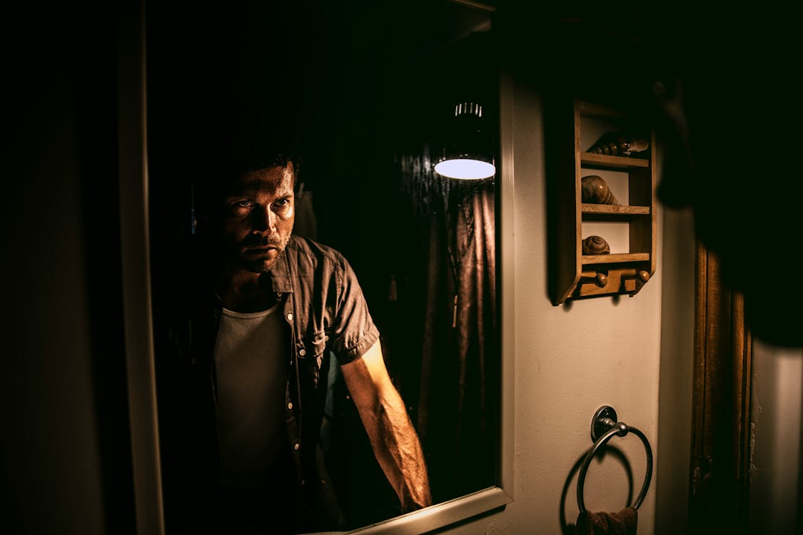 Free A man with a serious expression stands in front of a mirror in a dimly lit room. Stock Photo Free A man with a serious expression stands in front of a mirror in a dimly lit room. Stock Photo