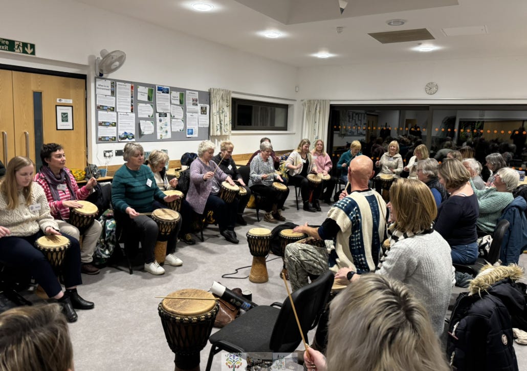A group of people sit in a circle in a bright community room, each holding a djembe drum. They appear to be taking part in a group drumming session, focused and engaged. The room has white walls, large windows with curtains, and a bulletin board covered with posters and notices. The atmosphere feels communal, lively, and collaborative.