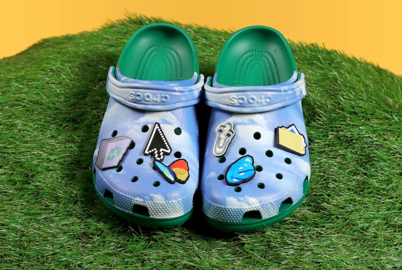 A pair of Crocs sitting on a hill of green turf; the Crocs feature recycling bin, Clippy, MSN, folder, Internet Explorer, and pointer jibbitz