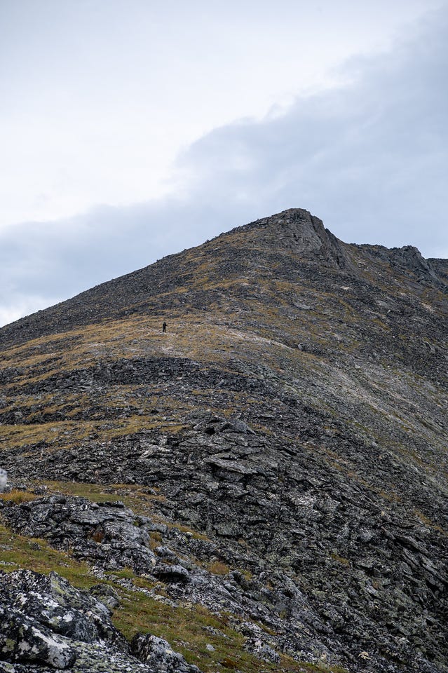 A tiny lone figure stands partway up an enormous sweep of grey scree and talus that climbs to a rounded rocky summit, the vast slope dwarfing the hiker under an overcast sky.