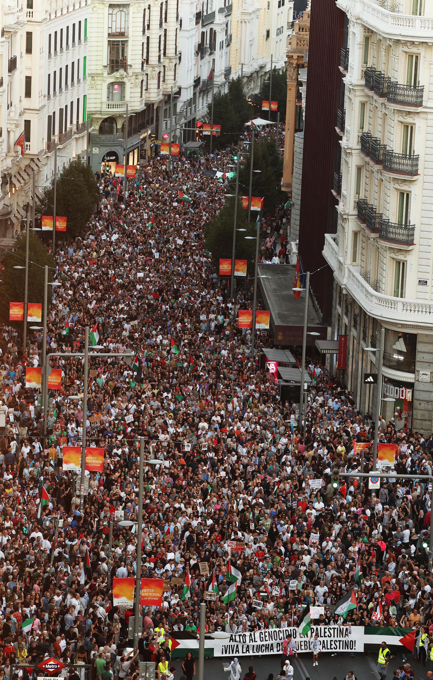 Una veduta aerea inquadra una strada cittadina gremita di persone che partecipano a una grande manifestazione. La folla si estende lungo l'ampia strada, fiancheggiata da alti edifici con facciate chiare e finestre a balcone. I manifestanti sventolano bandiere palestinesi (verdi, bianche, nere e rosse) e tengono in mano cartelli. Striscioni arancioni con scritte sono appesi ai lampioni lungo la strada. Una veduta aerea inquadra una strada cittadina gremita di persone che partecipano a una grande manifestazione. La folla si estende lungo l'ampia strada, fiancheggiata da alti edifici con facciate chiare e finestre a balcone. I manifestanti sventolano bandiere palestinesi (verdi, bianche, nere e rosse) e tengono in mano cartelli. Striscioni arancioni con scritte sono appesi ai lampioni lungo la strada.