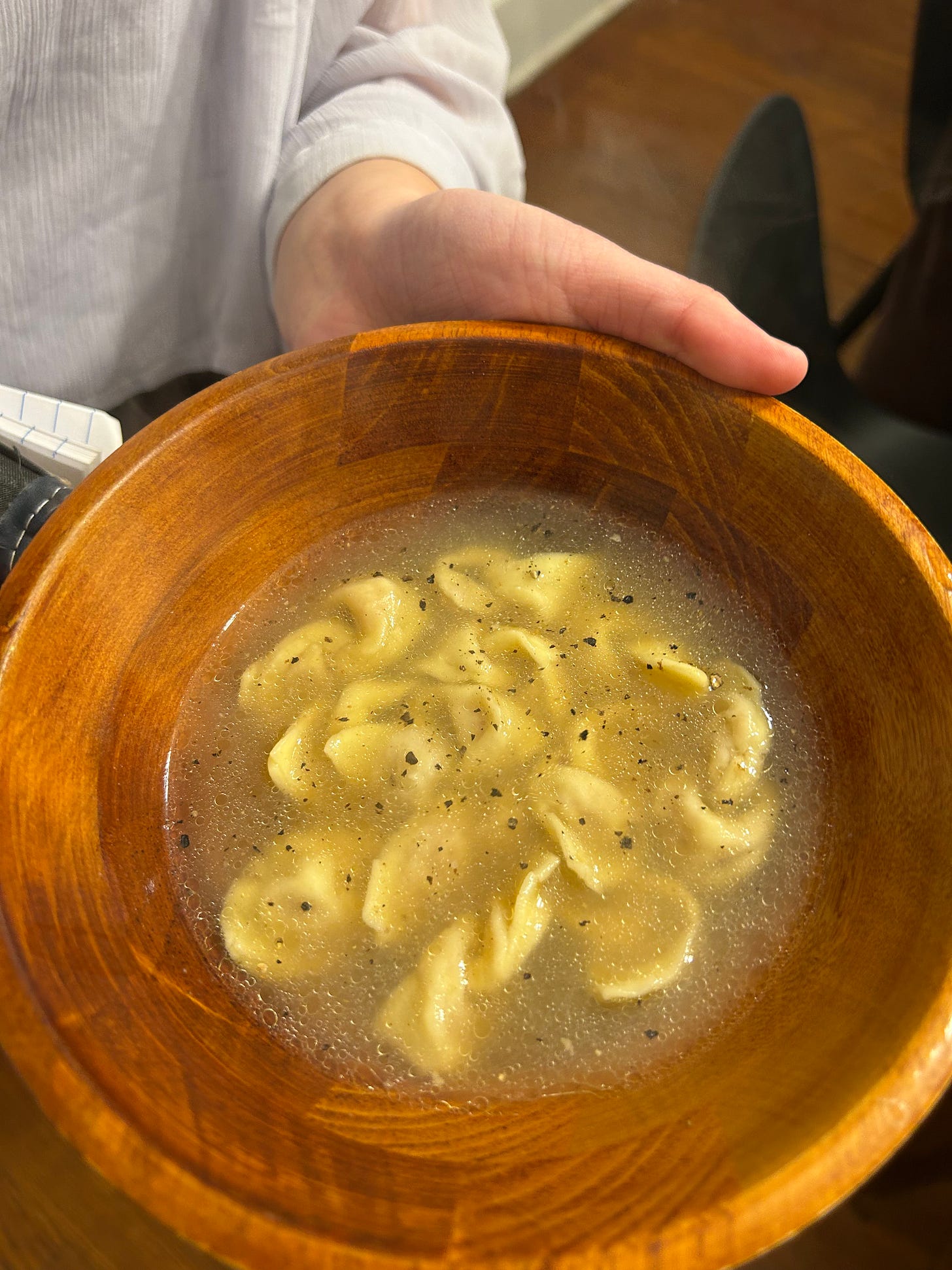 A hand holding a brown bowl of tortellini in broth.