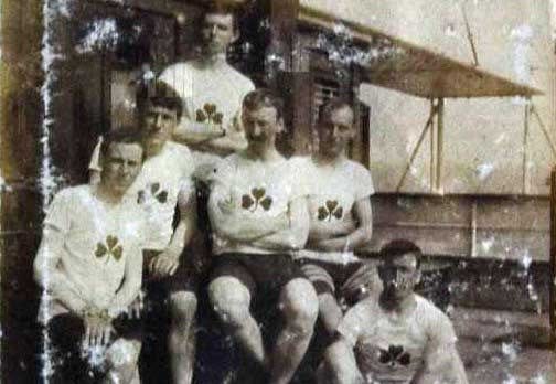 A grainy, black-and-white photograph of the 1912 Irish Olympic Cycling Team. Six men are posing together, wearing white jerseys adorned with a green shamrock emblem on the chest. The athletes, who appear strong and determined, are seated and standing in front of a wooden structure, possibly aboard a ship en route to the Stockholm Olympics. The image shows signs of age, with scratches and faded areas, but the pride and camaraderie of the team remain visible.
