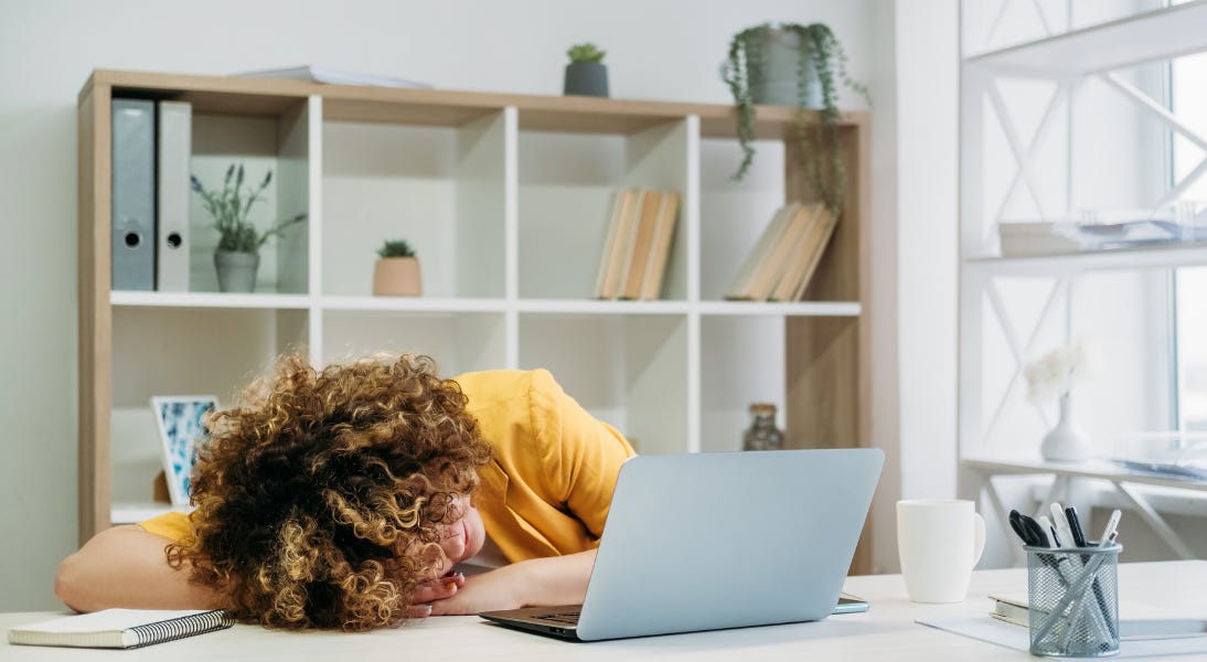 Woman at a desk with a computer in a professional setting with her head down indicating she's tired and struggling to focus.