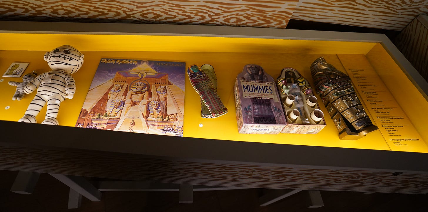 A display case in the Paris Momies exhibition containing mummy-themed toys and games, including a stuffed mummy and boxed games.