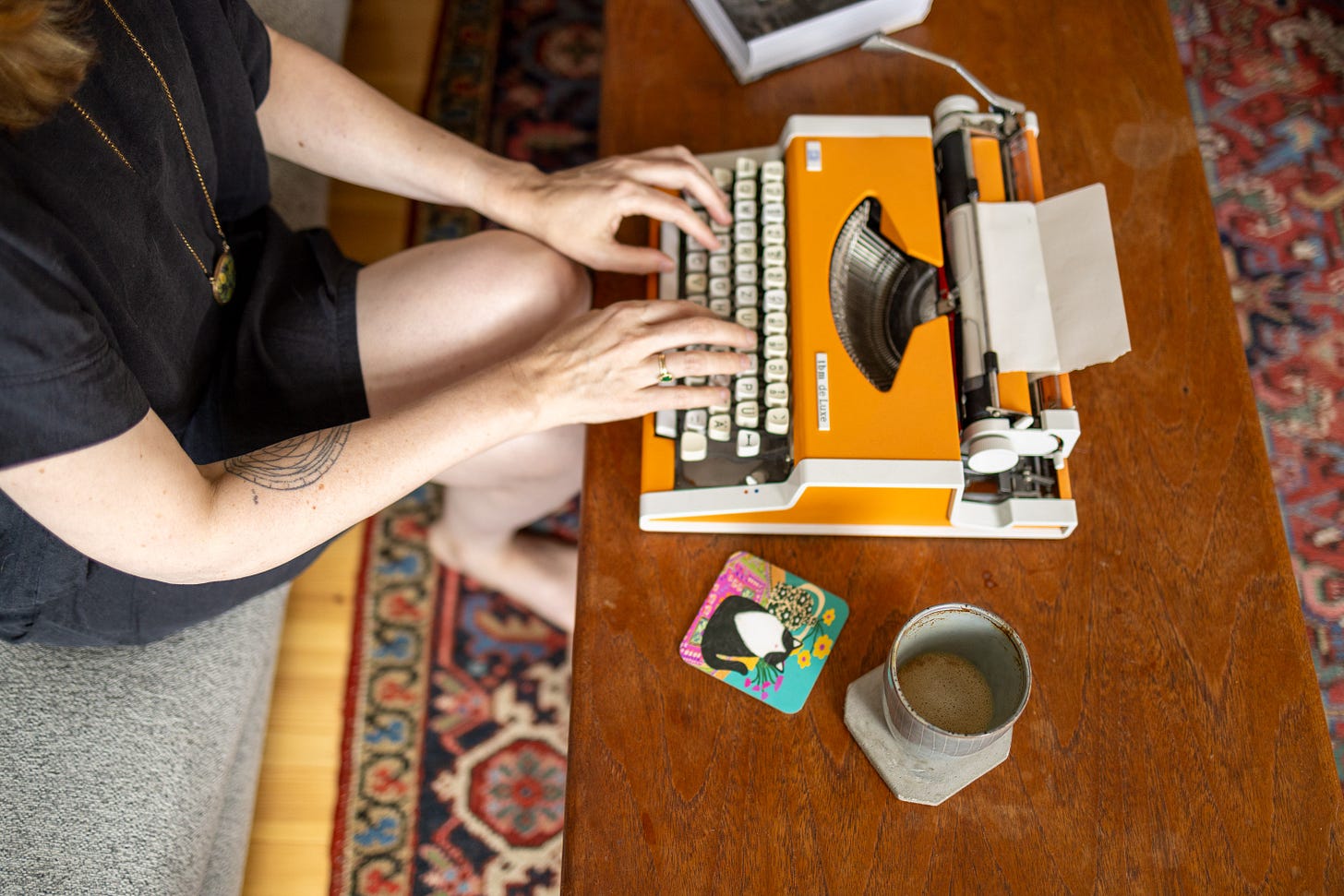 A white woman in a short-sleeved black dress sits on a sofa and types on the orange typewriter in front of her with a cup of coffee next to the typewriter